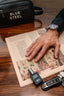 Hand with tattoo holding a 'Blue Steel' bottle on a wooden surface with newspaper and keychain.