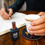 Person holding a cup with a book and perfume bottle on a wooden table