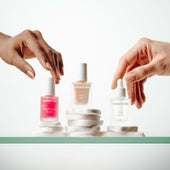 Three bottles of NONICURE nail and cuticle care products on a white surface with hands reaching towards them.